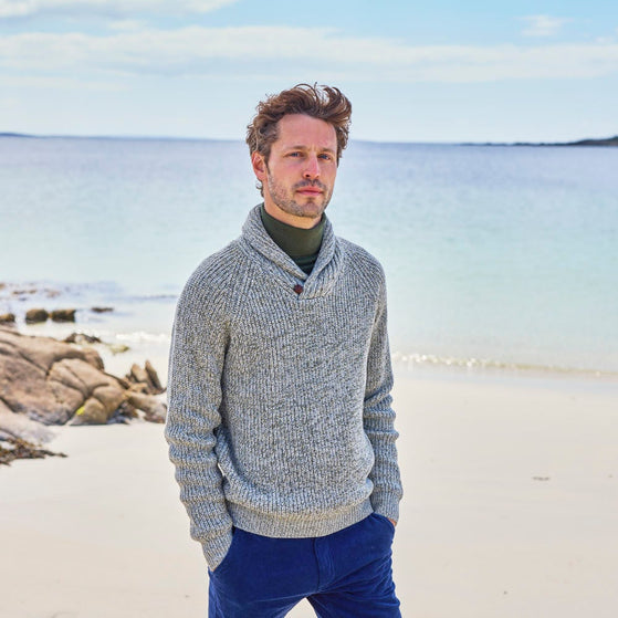 A man standing on a beach wearing a gray shawl collar button sweater and blue trousers.