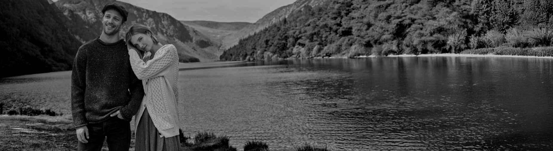 Black and white photo of a couple wearing traditional Irish aran sweaters embracing by a lake with mountains in the background