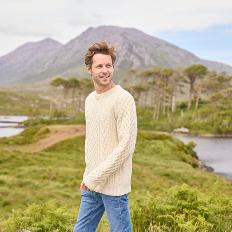 A man standing outdoors wearing a beige hand knit Aran sweater with a mountain and trees in the background.