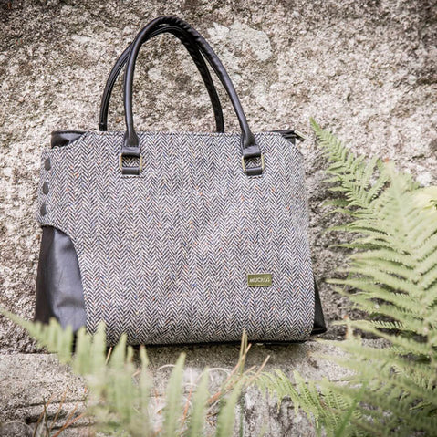 A handbag with a tweed body and vegan leather details, placed on a stone surface with foliage in the background.