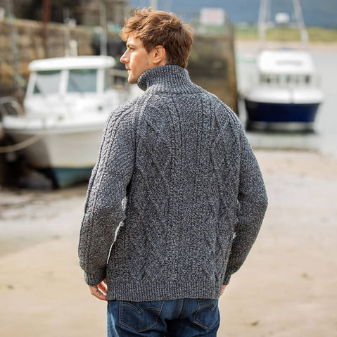 A man stands on a sandy beach, viewing boats moored in the background. He is wearing a textured gray aran sweater with a high collar, facing away from the camera. The setting suggests a coastal environment with calm water and rocky areas.