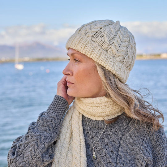 A woman wearing a cream-colored aran cable scarf and hat, with a knit pattern, standing by a body of water.