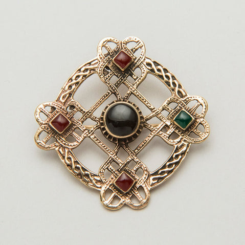An ornate Celtic brooch featuring a cross design with interwoven patterns. It has four decorative squares, each containing a small gemstone: two red and one green, with a central black stone. The piece combines intricate metalwork with colored accents.