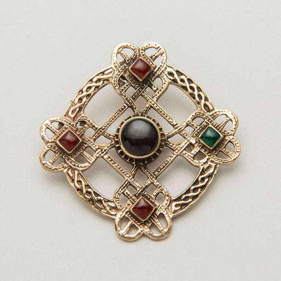 An ornate Celtic brooch featuring a cross design with interwoven patterns. It has four decorative squares, each containing a small gemstone: two red and one green, with a central black stone. The piece combines intricate metalwork with colored accents.