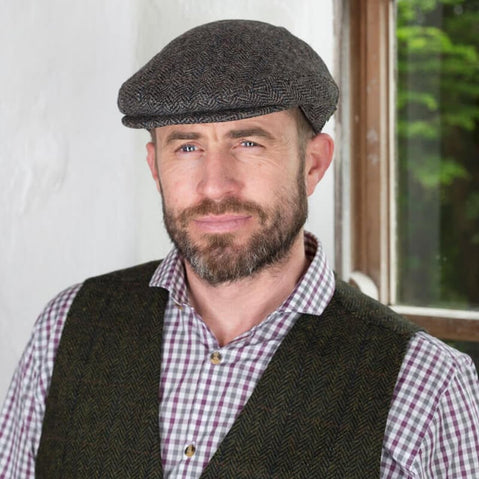 A man with a beard and wearing an Irish tweed flat cap stands in front of a window. He is dressed in a checked shirt and a dark vest, with a neutral expression. The background features natural light coming through the window, highlighting the texture of the wall.