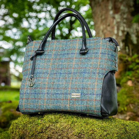 A handbag with a tweed body and vegan leather details, placed on a stone surface with foliage in the background.
