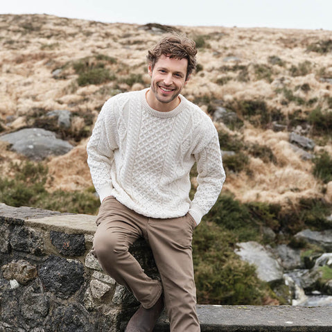 A man wearing a cream fisherman knit sweater with a crew neck, sitting on a stone wall in a landscape with dry grass and a body of water in the background.