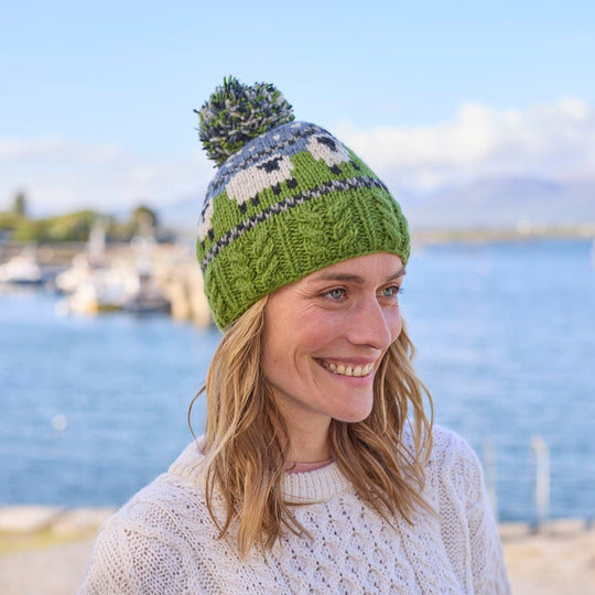 A woman wearing a blue and green bobble hat with a sheep design and aran cable stitching, standing in front of a body of water.