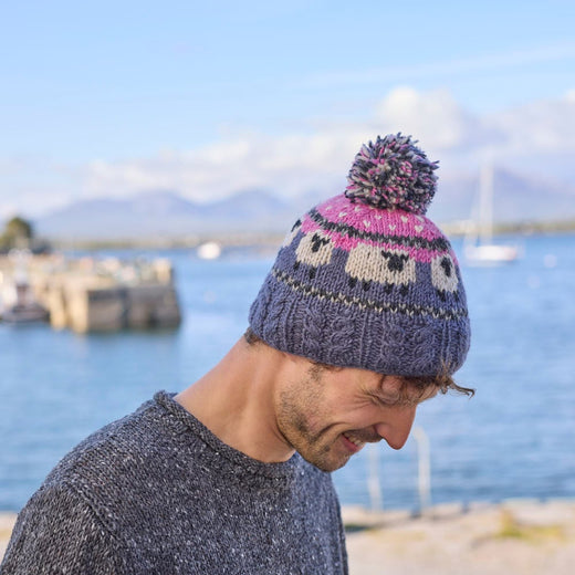 A person wearing a knitted bobble hat with a sheep design in pink and blue colors on a grey background.