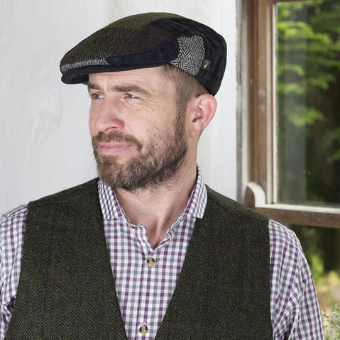 A man with a beard wearing an Irish tweed flat cap and a checked shirt under a dark vest stands near a window, looking thoughtfully to the side. Natural light illuminates the scene, highlighting the indoor setting.