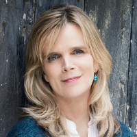 Lulu O'Sullivan, Founder & CEO of The Irish Store, is a woman with wavy, shoulder-length blonde hair is smiling gently at the camera, wearing a blue top and white shirt. The background features a weathered wooden surface, suggesting an outdoor or rustic setting.