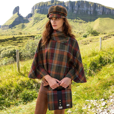 A person standing in a field wearing a brown, green, and red tweed poncho with a matching tweed cap.