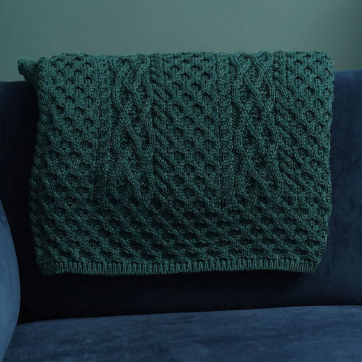 A textured, green knitted Aran blanket drapes over a dark blue couch in a The wall behind is painted a muted green, complementing the overall warm aesthetic.