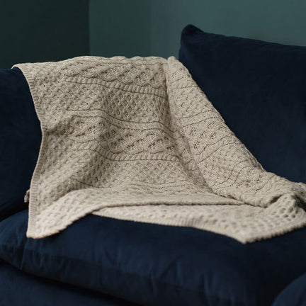 A textured, beige knitted Aran blanket drapes over a dark blue couch in a cozy setting. The wall behind is painted a muted green, complementing the overall warm aesthetic.