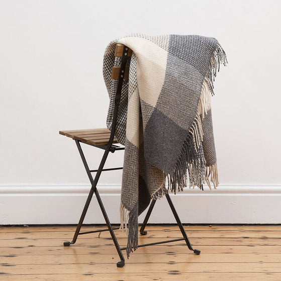 A checkered black, white, and gray lambswool/cashmere herringbone blanket draped over a chair against a white wall and wooden floor background.
