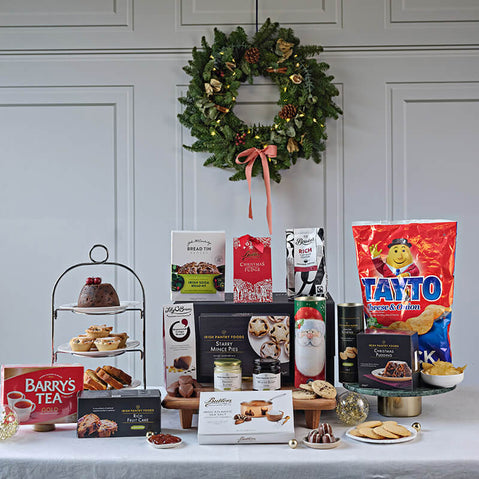 Traditional Irish Feast Hamper. A festive display featuring an assortment of food items arranged on a table. In the foreground, there's a multi-tiered stand with baked goods and treats. Several boxes of tea and snacks are visible, including Barry's Tea and Tayto crisps. At the back, a green wreath adorned with red accents hangs on the wall, adding to the holiday theme.