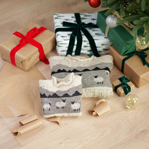 Gifts wrapped in brown paper with red and green ribbons, and a knitted sweater with sheep pattern on a wooden floor.