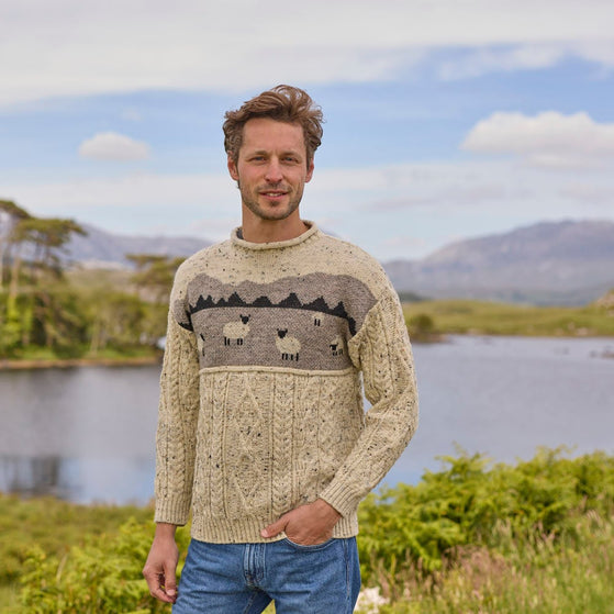 A man standing outdoors wearing a beige Aran sweater with a sheep landscape design.
