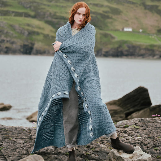 Image featuring the Aran Sheep Throw in blue, crafted with authentic Irish Aran cable knit patterns and a distinctive sheep motif border, showcasing traditional Irish craftsmanship in a scenic coastal setting.