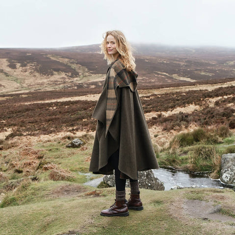 A woman stands on a grassy hill, wearing a dark green walking cape and brown boots. She faces slightly to the side, with a gentle smile. The background features rolling hills and heather under a cloudy sky. A small stream flows nearby, adding to the serene landscape.