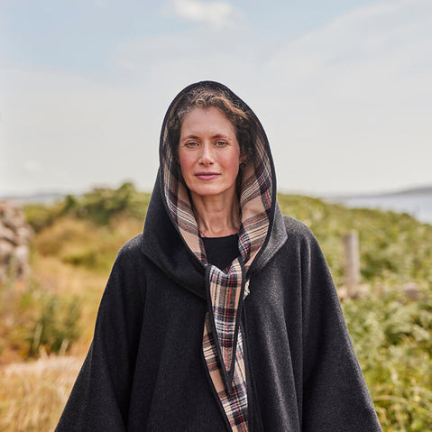 A woman stands outdoors, wearing a dark grey hooded cape with a plaid scarf. She has curly hair and a neutral expression. The background features green grass and a body of water under a clear sky.