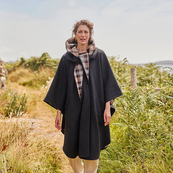 A woman stands on a rural path, wearing a dark grey cape with a hood lined in plaid. She has curly hair and is surrounded by greenery and tall grass, suggesting a serene outdoor setting. The background features a gentle landscape under clear skies.