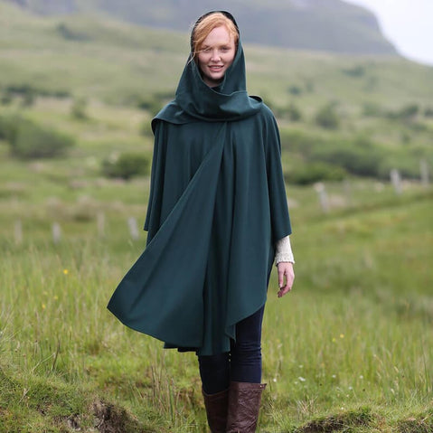 A woman stands in a grassy field, wearing a long, dark green cape with a hood. She has light brown hair and is smiling softly. The background features rolling hills and lush greenery, indicating an outdoor setting.