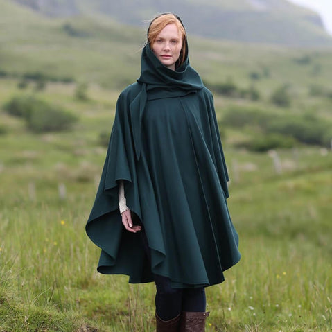 A woman stands in a grassy landscape, wearing a dark green cape with a hood. The background features rolling hills under a cloudy sky. Her expression is serene and she appears to be enjoying the natural surroundings.