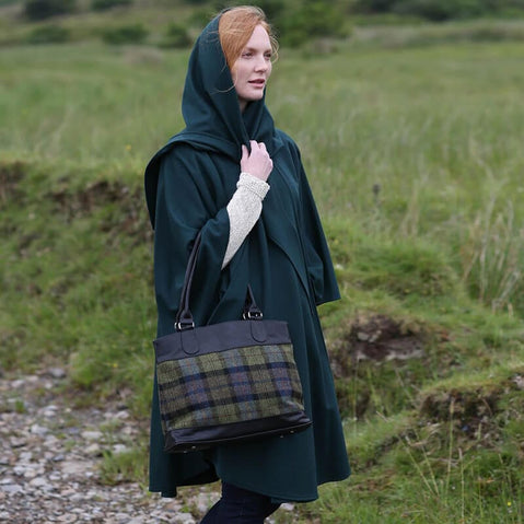 A woman stands in a grassy landscape wearing a dark green cape and a hood. She carries a plaid tweed handbag and looks off into the distance. The setting appears serene, with rolling hills in the background.