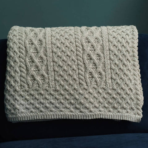 A textured, beige knitted Aran blanket drapes over a dark blue couch in a The wall behind is painted a muted green, complementing the overall warm aesthetic.