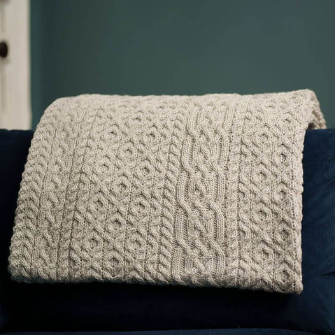 A silver marl merino aran throw with a geometric pattern draped over a dark-colored sofa cushion.