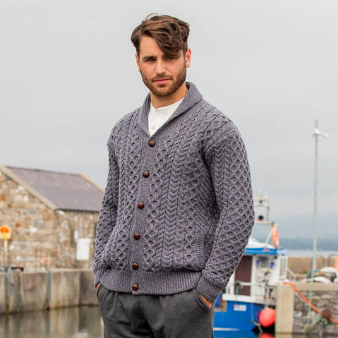 A man wearing a gray Aran cardigan with a high shawl collar and traditional brown leather buttons, standing in front of a harbor scene.