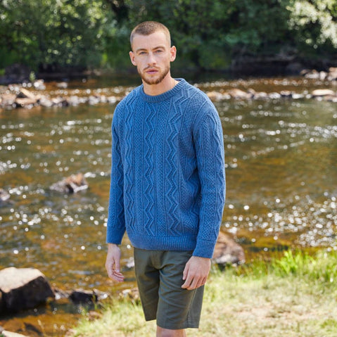 A man wearing a blue crew neck Aran sweater with a geometric pattern, standing beside a river.