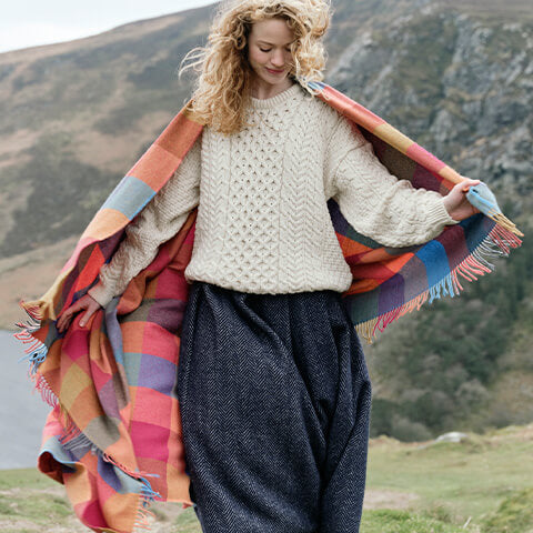 Woman wearing a traditional Aran sweater and draped in a colorful Irish wool check throw blanket from the homeware collection.
