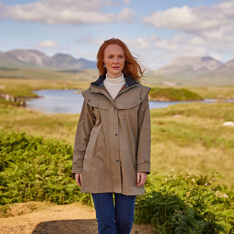 A woman in a light brown waterproof jacket stands on a grassy landscape with mountains and a body of water in the background. She wears a white turtleneck and blue jeans, looking directly at the camera against a setting that suggests outdoor exploration.