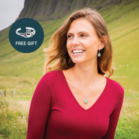 A woman with long, wavy hair smiles while wearing a red top and matching celtic earrings and necklace. She stands outdoors against a backdrop of green hills. A graphic in the corner indicates a promotional