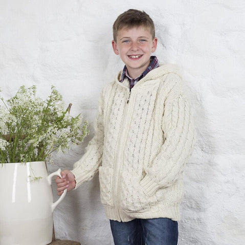 A smiling boy stands beside a large white vase filled with greenery. He wears a cozy cream-colored knit aran sweater with a hood and has a checkered shirt underneath. The background features a textured white wall.