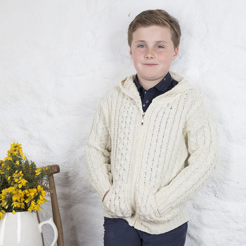 A young boy stands confidently in a cozy, cream-colored knitted aran sweater with a hood. He is wearing a dark shirt underneath and has his hands in the pockets of the sweater. The background features a textured white wall, and there is a wooden chair beside him with a vase of yellow flowers.