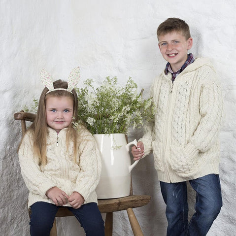 A young boy and girl pose together in a cozy setting. The boy stands beside a white watering can filled with flowers, wearing a beige knitted aran sweater. The girl sits on a wooden chair, also in a beige aran sweater, with bunny ear headbands adorning her hair. The background features a light-colored stone wall.