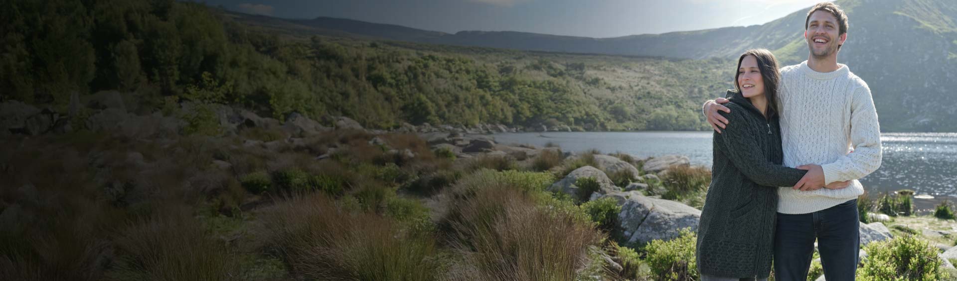 Two people standing together in a scenic outdoor setting with mountains and water wearting authentic lightweight aran sweater and cardigan