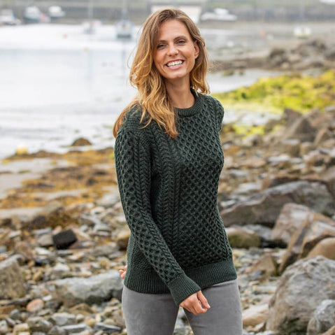 A person wearing a Women's Traditional Crew Neck Aran Sweater in army green stands outdoors on a rocky shoreline with the sea and boats in the background. The sweater features intricate cable knit patterns.