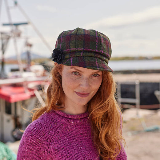 A woman with long red hair smiles while wearing a plaid cap and a knitted purple sweater. She is standing outdoors near a harbor, with boats visible in the background under a bright sky.