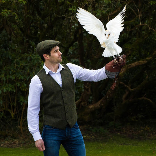 A person wearing a green Irish wool Trinity flat cap holding a white owl on their extended hand, outdoors with trees in the background.
