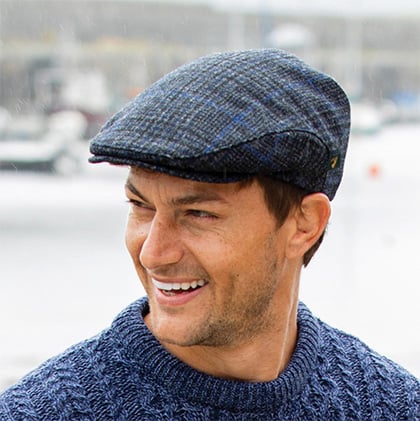 A man is smiling while wearing a dark blue flat cap and a knit sweater. He is outdoors, near a body of water, with boats visible in the background. The atmosphere appears overcast and slightly rainy.