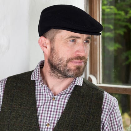 A man with a beard wearing a flat cap and a patterned shirt stands near a window, looking thoughtfully to the side. He is dressed in a dark vest over the shirt, with greenery visible outside.