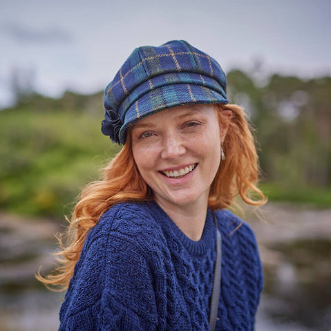 A woman with long red hair smiles while wearing a blue plaid hat and a textured blue sweater. She is outdoors, with greenery blurred in the background. The scene suggests a relaxed and cheerful atmosphere.