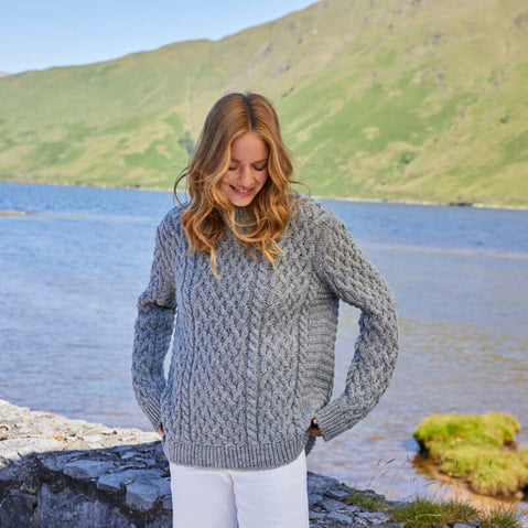 A woman standing by a lake wearing a gray Aran sweater with a ribbed turtle neck and cuffs.