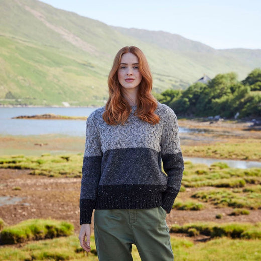 A woman standing in a scenic location wearing a color-blocked Aran sweater with a wide neck and a slim fit.