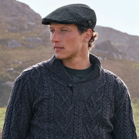 Men's Heritage Plaid Irish Flat Cap Green Check with traditional Irish Aran sweater in a mountain landscape.