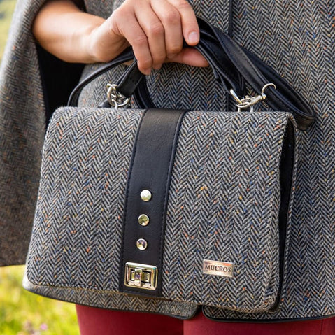 Fiona tweed handbag with black leather trim and handles, featuring a herringbone pattern and metal clasp, held at the side of a person wearing a matching tweed garment.